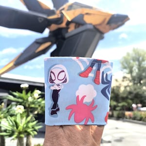 Pode incluir: Capa de copo de café azul claro com um padrão repetido de Spider-Gwen e outros personagens do Homem-Aranha. A capa apresenta personagens de desenhos animados, nuvens e desenhos em forma de estrela rosa. O fundo inclui uma grande estrutura amarela e preta.