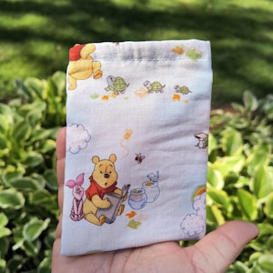 May include: A light blue fabric pouch featuring illustrations of Winnie the Pooh, Piglet, turtles, and honey pots. The pouch is held in a hand against a backdrop of green foliage.