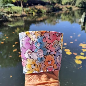 Pode incluir: Uma manga de tecido para copo com um padrão repetitivo de ursos de desenho animado coloridos em tons de rosa, roxo, azul, amarelo e laranja. Cada urso exibe um símbolo único no peito, incluindo corações, luas e flores sobre um fundo claro.