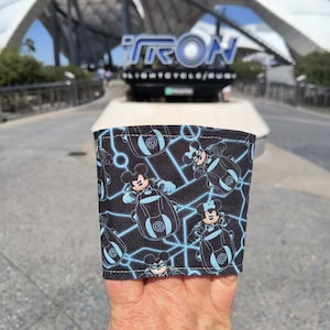 Pode incluir: Um bracelete de tecido preto e azul com um padrão repetido de Mickey Mouse pilotando uma moto de luz com a palavra "TRON" no fundo.
