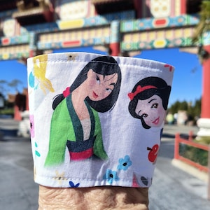 Pode incluir: Capa de copo de café em tecido branco com ilustrações de Mulan e Branca de Neve. A capa tem um padrão floral com detalhes em azul, amarelo e rosa. Um arco colorido está no fundo.