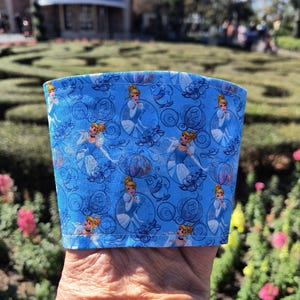 以下が含まれることがあります： 青い生地のコーヒーカップスリーブ。シンデレラが舞踏会用のドレスとカボチャの馬車に乗っている模様が繰り返されています。スリーブは持ち上げられ、緑と建物のあるぼやけた屋外の背景に対してデザインが示されています。