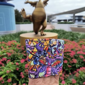 Peut inclure: Un manchon de boisson en tissu coloré avec un motif répété du dragon violet Figment de l'attraction Disney. Le manchon est tenu devant une statue de dragon en bronze et des fleurs roses.