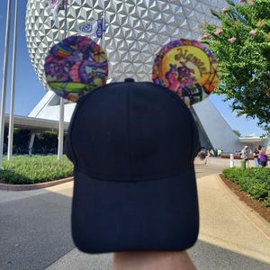 Puede incluir: Una gorra de béisbol negra lisa con dos orejas circulares unidas en la parte superior. La oreja izquierda muestra un gráfico colorido de un dragón morado, mientras que la derecha presenta un dragón morado con el texto Figment en amarillo.