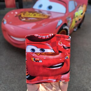 Pode incluir: Boneco de dedo vermelho com uma imagem em close-up de Relâmpago McQueen do filme Cars da Disney Pixar. O boneco tem um padrão xadrez e o texto "Kachow!" no lado.