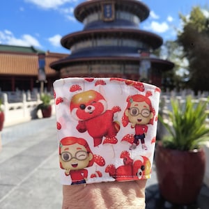 May include: White fabric coffee cup sleeve featuring a repeating pattern of a red panda character and red mushrooms. The red panda is depicted in various poses. The background includes a blurred view of a blue-roofed building and a sunny sky.
