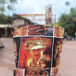 Może przedstawiać: Materiałowa osłonka na kubek do kawy z kolażem plakatów filmowych Indiana Jones. Wzór zawiera tytuły Indiana Jones and the Temple of Doom oraz Indiana Jones and the Raiders of the Lost Ark, wraz z wizerunkiem głównego bohatera.