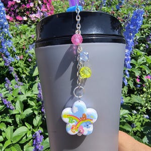 Peut inclure: Un gobelet gris avec un couvercle noir et une paille violette. Une chaîne décorative avec des perles roses, bleues et jaunes est suspendue au couvercle, attachée à un charm en forme de fleur représentant les personnages des Care Bears et un arc-en-ciel.