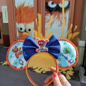 May include: A pair of orange and blue Mickey Mouse ears with a blue bow. The ears are decorated with images of characters from the Muppets, including Cookie Monster, Elmo, and Kermit the Frog. The ears are in front of a sign that says "The Muppets".