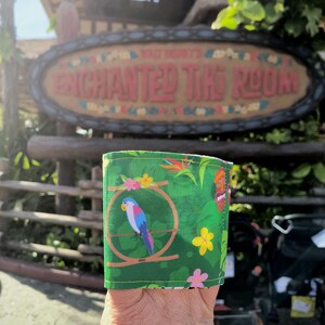 May include: A green fabric item with a tropical parrot design. The parrot is inside a circular cage, surrounded by flowers and leaves. The background features the sign for the Enchanted Tiki Room.