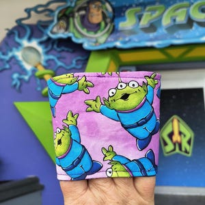 May include: A purple and blue fabric drink sleeve featuring a repeating pattern of green aliens in blue space suits. The aliens have three eyes and are in various poses. The background is a light purple color.
