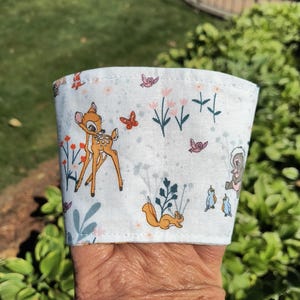 Pode incluir: Manga de xícara de café de tecido branco com uma cena florestal extravagante com Bambi, um esquilo, pássaros e borboletas. O design inclui flores cor-de-rosa e folhagem verde, criando uma estética charmosa inspirada na natureza.