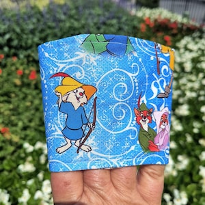 Może przedstawiać: Niebieski rękaw z tkaniny z postaciami z kreskówki z filmu "Robin Hood". Rękaw ma jasnoniebieskie tło z białymi wirami i płatkami śniegu. Postacie obejmują królika z łukiem i strzałą.