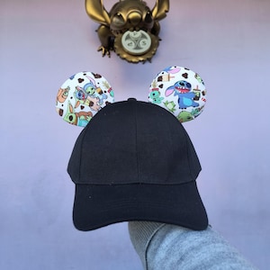 May include: Black baseball cap with Stitch character ear covers. The ear covers are white with a colorful print of Stitch characters.