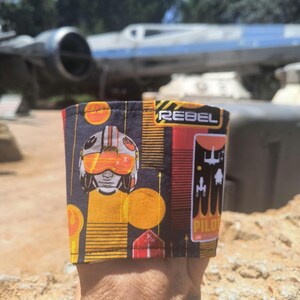 May include: A fabric cup sleeve featuring a black background with a repeating pattern of orange circles, yellow lines, and a pilot helmet graphic. The text REBEL and PILOT are printed in white and black. The design includes stylised starfighter silhouettes.