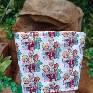 Pode incluir: Manga de copo de café de tecido branco com um padrão repetido de mulheres de desenho animado. As mulheres estão em várias roupas e penteados, segurando bolos. O fundo é um exuberante jardim verde.