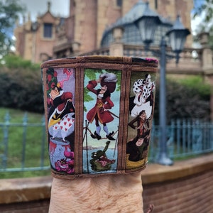 Può includere: Una custodia in tessuto per tazza con illustrazioni incorniciate di cattivi Disney. La custodia presenta la Regina di Cuori, Capitan Uncino e Crudelia De Mon, con un bordo marrone. Lo sfondo include una vista sfocata di un edificio.