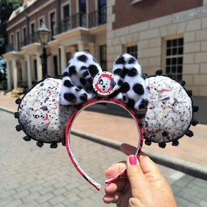 May include: A headband featuring two circular ears covered in a white fabric with a black dalmatian puppy print and black pom-pom trim. A central bow made of black and white faux fur sits between the ears, accented by a circular dalmatian puppy emblem.