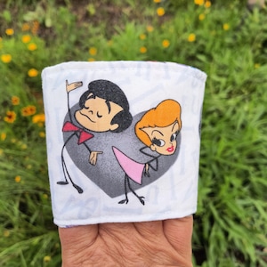 Pode incluir: Uma manga de tecido branco para copo com uma ilustração de desenho animado de um homem de cabelo preto com gravata borboleta vermelha e uma mulher de cabelo laranja com saia rosa, de costas um para o outro diante de um coração cinza.
