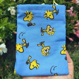 Könnte beinhalten: Ein hellblauer Stoffbeutel mit einem sich wiederholenden Muster der gelben Woodstock-Figur aus den Peanuts-Comics. Die kleine rechteckige Tasche hat eine genähte Oberkante und enthält dünne gelbe Kordelzüge an der oberen Öffnung.
