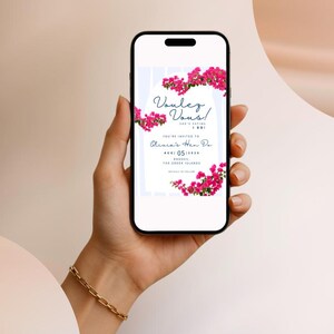 May include: A smartphone displays a digital invitation with the text "Voulez Vous!" and "Olivia's Hen Do". The invitation is decorated with pink flowers. The phone is held in a hand with a gold bracelet.