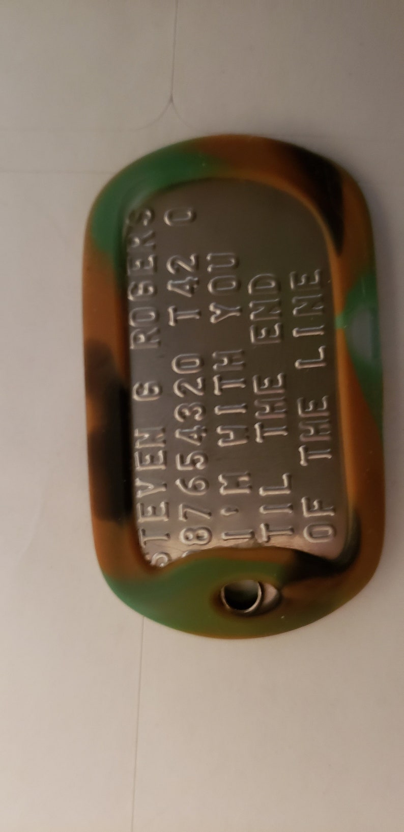May include: A camouflage-colored dog tag with the text "STEVEN G. ROGERS  87654320  I'M WITH YOU  'TIL THE END  OF THE LINE" engraved on a silver metal plate.