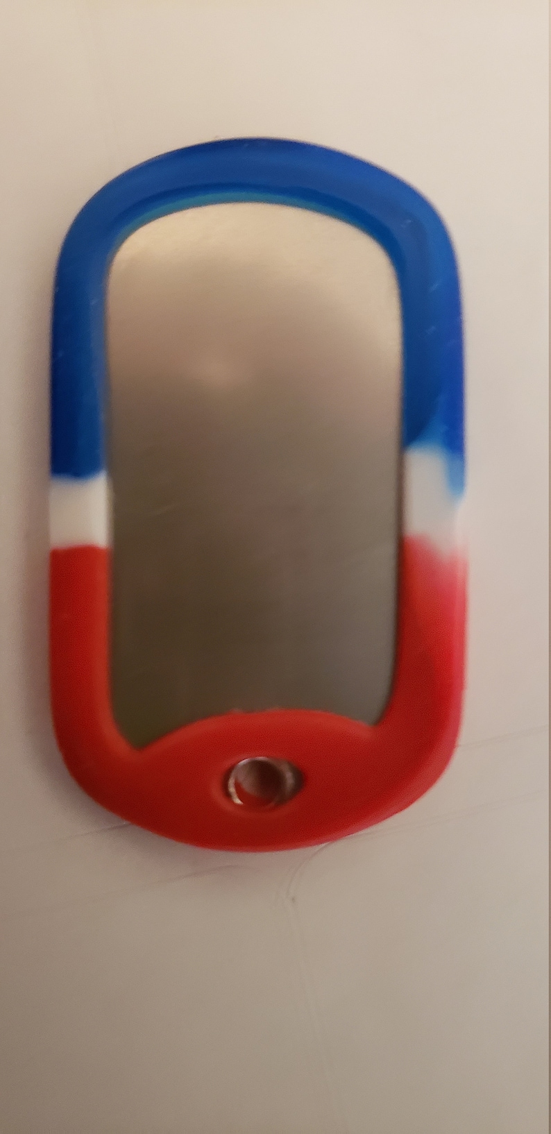 May include: A red, white, and blue silicone dog tag with a silver metal insert. The tag has a small hole at the top for attaching a chain or lanyard.