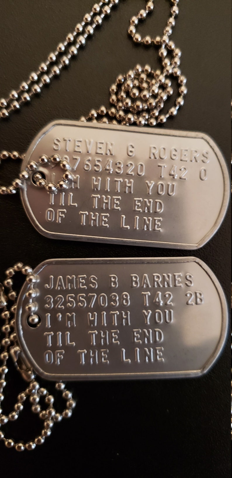 May include: Two silver dog tags on a silver chain. The top tag reads "STEVEN G ROGERS 87654320 T42 0 I'M WITH YOU TIL THE END OF THE LINE". The bottom tag reads "JAMES B BARNES 32557038 T42 2B I'M WITH YOU TIL THE END OF THE LINE".