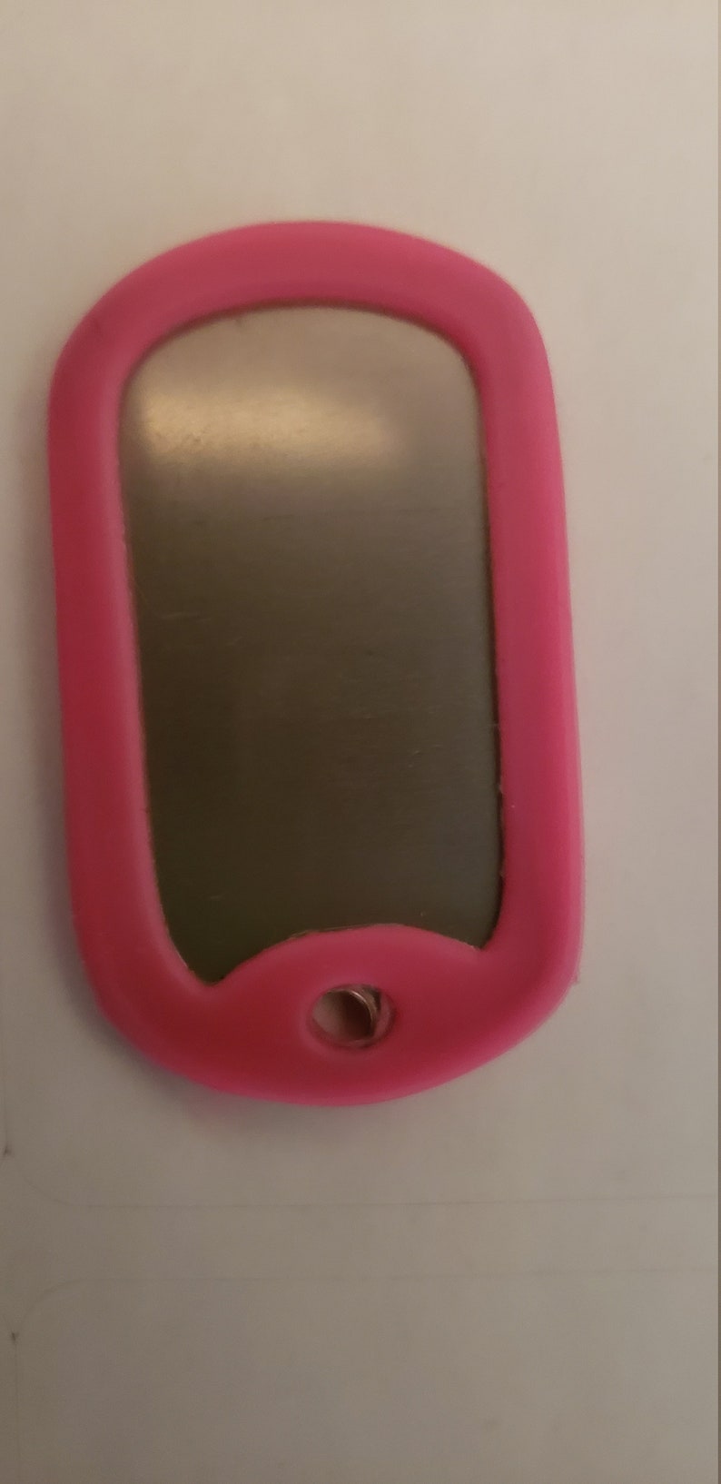 May include: A pink silicone dog tag with a silver metal insert. The tag has a hole at the top for attaching a chain or lanyard.