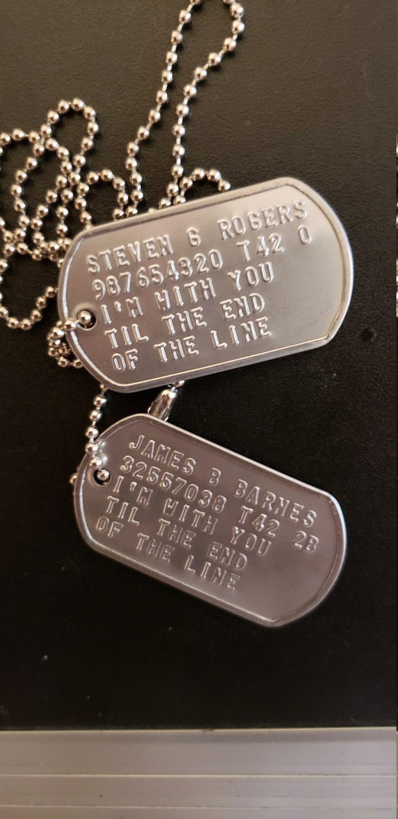 May include: Two silver military dog tags on a silver chain. The top tag reads "STEVEN 6 ROGERS 987654320 T42 0 I'M WITH YOU TIL THE END OF THE LINE". The bottom tag reads "JAMES B BARNES 32557038 T42 28 I'M WITH YOU TIL THE END OF THE LINE".