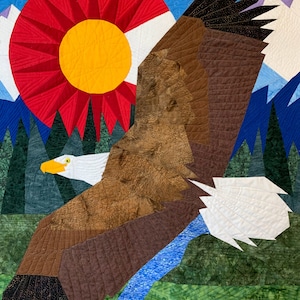 Op de afbeelding: Een quilt die een Amerikaanse zeearend toont die over een bergketen vliegt met een rode, witte en gele zon op de achtergrond. De vleugels van de adelaar zijn wijd uitgespreid en zijn klauwen zijn uitgestrekt. De bergen zijn groen en blauw, en de zon schijnt helder.