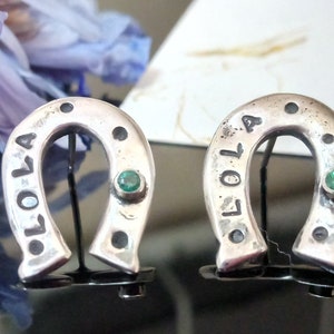 May include: A pair of silver horseshoe earrings with green gemstones. The earrings are engraved with the word "Lola" on one side and "2014" on the other.