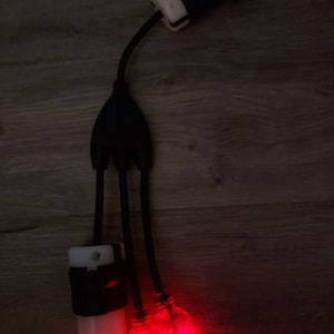 May include: A black electrical power splitter cable featuring one input plug and three output connectors. One output is a white cylindrical connector, while the other two are transparent red illuminated connectors that glow brightly when powered.