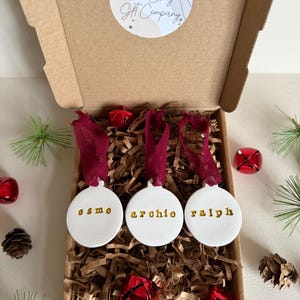 Op de afbeelding: Een open kartonnen doos met drie witte, ronde ornamenten met gouden namen: "esme", "archie" en "ralph". Ze hangen aan bordeauxrode linten. De doos is gevuld met bruin papier, met dennentakken en rode belletjes.