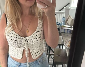 Op de afbeelding: Crèmekleurige gehaakte crop top met een striksluiting, gecombineerd met lichtblauwe denim jeans. De top heeft een delicaat open weefselontwerp. De jeans hebben een klassiek ontwerp met vijf zakken en distressed details. De outfit is vastgelegd in een spiegel selfie.