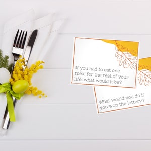 Two conversation starter cards sit on a white table next to a fork and knife decorated for Thanksgiving.