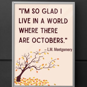 Tan colored poster with a silver frame against a black wall. The poster is fall-themed with a picture of a tree losing orange and brown leaves. It has a quote in block letters, Im so glad I live in a world where there are Octobers. LM Montgomery