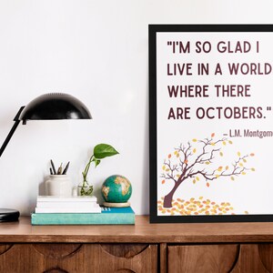 Picture of a desk with a light and books next to a black framed poster leaning against the wall. The poster is fall themed with a picture of a tree losing its leaves and a quote that reads, Im so glad I live in a world where there are Octobers.
