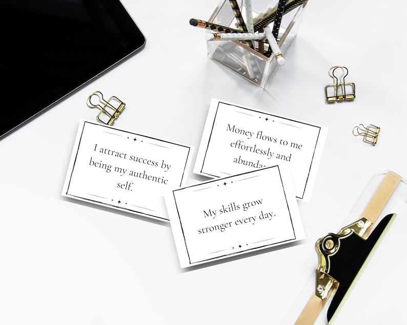 Three daily mantra cards are shown on a white desk with a clipboard, clips, and a container of pencils.