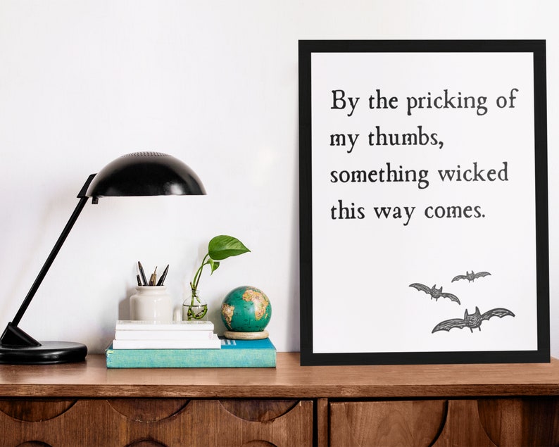 By the pricking of my thumbs, something wicked this way comes. Halloween art print sits on a desk next to lamps and book pile. This spooky literary quote art print is effortless Halloween decoration for any home office or classroom.