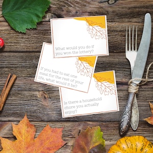 Thanksgiving conversation starters lay on a place setting for a Thanksgiving dinner with fall leaves, pumpkins, and a fork and knife.