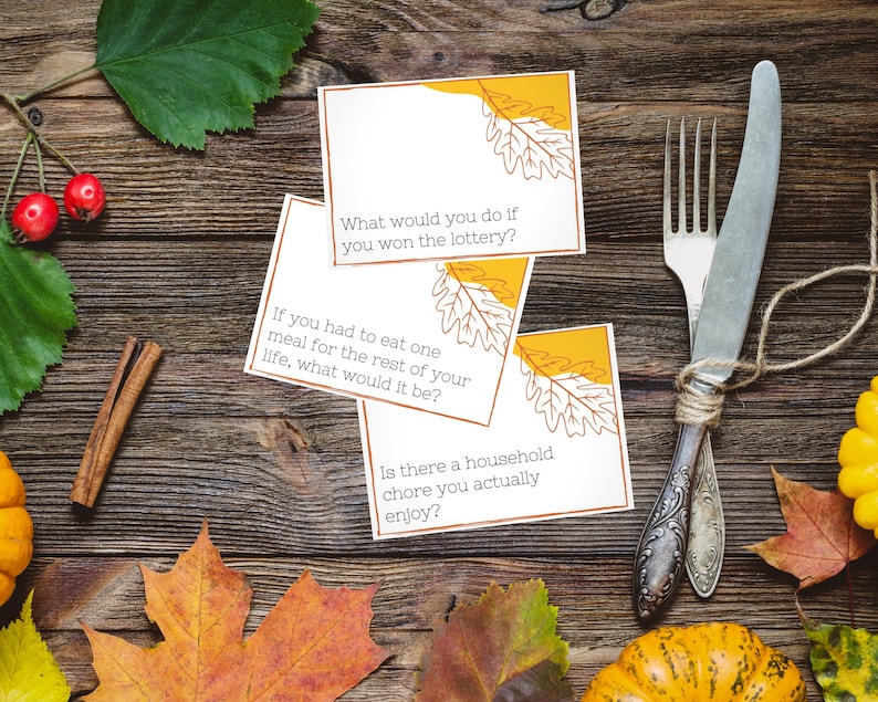Thanksgiving conversation starters lay on a place setting for a Thanksgiving dinner with fall leaves, pumpkins, and a fork and knife.
