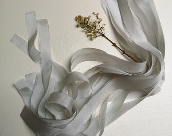 May include: A bundle of light gray satin ribbon, arranged in a loose, flowing pattern. The ribbon is soft and silky, with a subtle sheen.