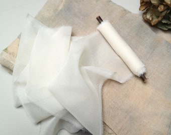 Op de afbeelding: Een wit zijden lint met rafelige randen, opgerold en vastgebonden met een bruine twijg, op een beige stof met een lichtblauw en grijs patroon.