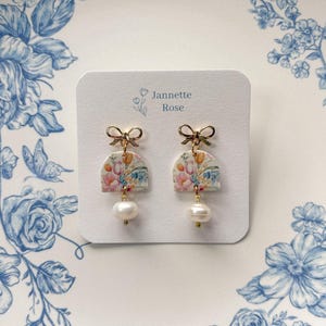 May include: Floral-patterned earrings with gold bow detail and a dangling pearl. The earrings are displayed on a white card with the text "Jannette Rose". The background features a blue floral design.
