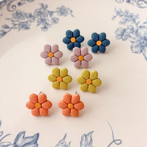 Könnte beinhalten: Eine Sammlung von blütenförmigen Ohrsteckern in verschiedenen Farben, darunter Blau, Rosa, Gelb und Orange. Jeder Ohrring hat eine kontrastierende Mitte. Die Ohrringe werden auf einem weißen Teller mit blauem Blumenmuster präsentiert.