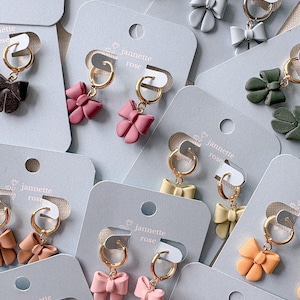 Puede incluir: Colección de pendientes con forma de lazo en varios colores, incluyendo rojo, rosa, beige y verde, exhibidos en tarjetas grises. Cada pendiente presenta un aro dorado. Las tarjetas llevan el texto "jannette rose".