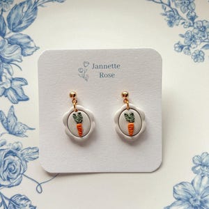 May include: A pair of white, scalloped-edge earrings with a gold post. Each earring features a small, round design with a carrot and green leaves. The earrings are displayed on a white card with the text "Jannette Rose".