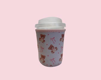 May include: A light blue neoprene cup sleeve featuring a repeating pattern of brown teddy bears with pink bows and small pink polka dots. The sleeve has a pink stitched trim at the top and bottom edges and fits around a clear plastic cup.