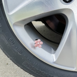 May include: A close-up of a silver car wheel with a pink flower-shaped valve stem cap. The tire has the text "DOT APOA OH9X 3619" printed on the side.